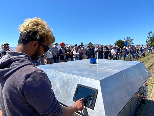 A young man hunches over a boxy machine in a field, as people stand on either side watching.