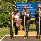 Four people dressed in suits and standing outside with shovels. Each one is smiling and has a foot up on one side of their shovel.